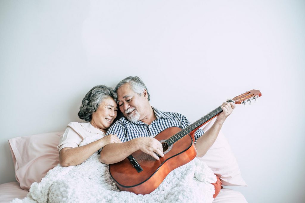 Älteres Paar liegt im Bett und der Mann spielt Gitarre